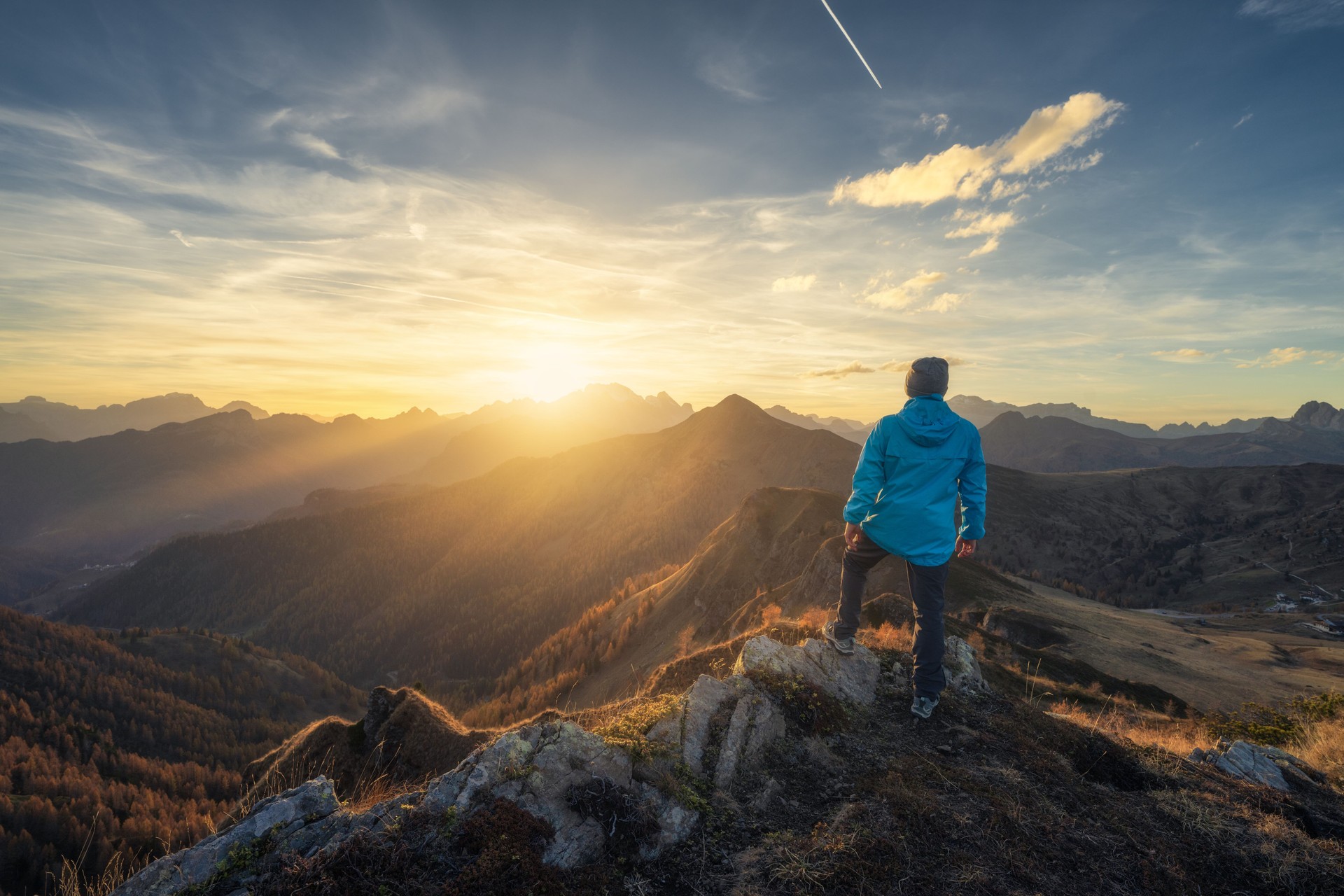 Man on stone on the hill and beautiful mountains in haze at colorful sunset in autumn. Dolomites, Italy. Sporty guy, mountain ridges in fog, orange grass and trees, blue sky with sun in fall. Hiking